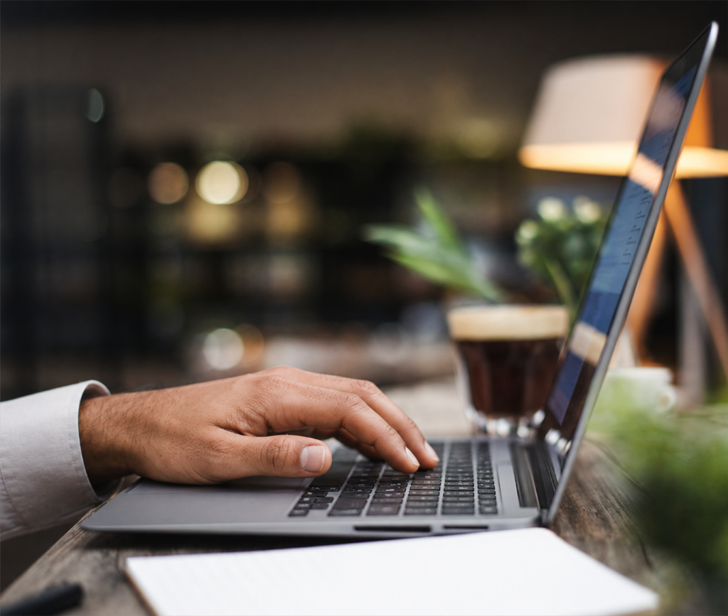 A man’s hand typing on a laptop keyboard at a cozy workspace, with a blurred coffee cup, plant, and warm lamp in the background.